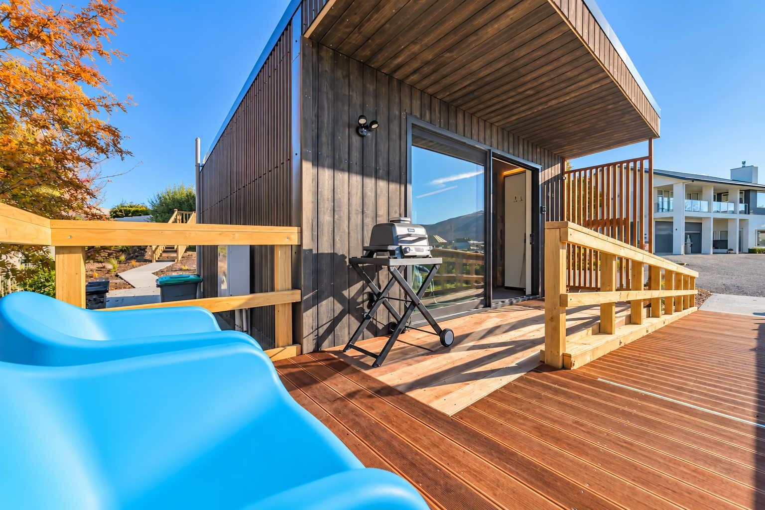 Una terraza de madera con una parrilla, un tobogán azul y una escalera de madera que lleva a un edificio con ventanas grandes.