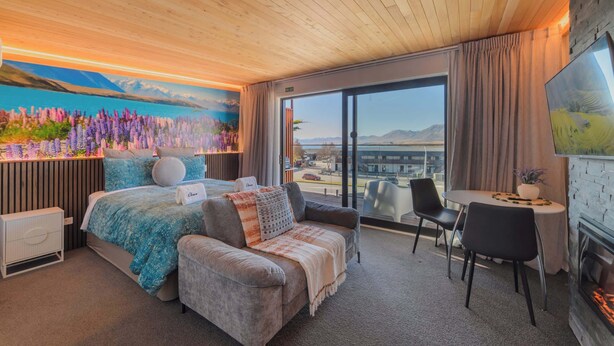 A hotel room with a bed, a sofa, a dining table with chairs, a fireplace, and a large window with a view of the mountains. - Aurora Vista - Ohana (Lake Tekapo)