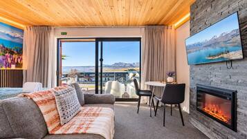 A hotel room with a wooden ceiling, a fireplace, a sofa with a patterned throw, a dining table with chairs, and a large window with a mountain view.