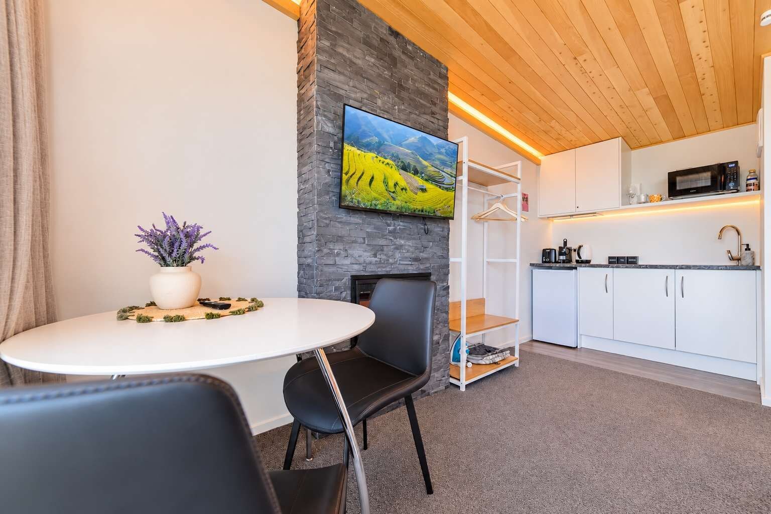 Une pièce moderne comprenant une table à manger, des chaises, une plante en pot, un téléviseur à écran plat fixé à un mur en pierre, un coin cuisine avec des placards et des appareils électroménagers, et un plafond en bois.