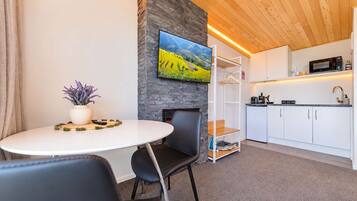 Une pièce moderne comprenant une table à manger, des chaises, une plante en pot, un téléviseur à écran plat fixé à un mur en pierre, un coin cuisine avec des placards et des appareils électroménagers, et un plafond en bois.
