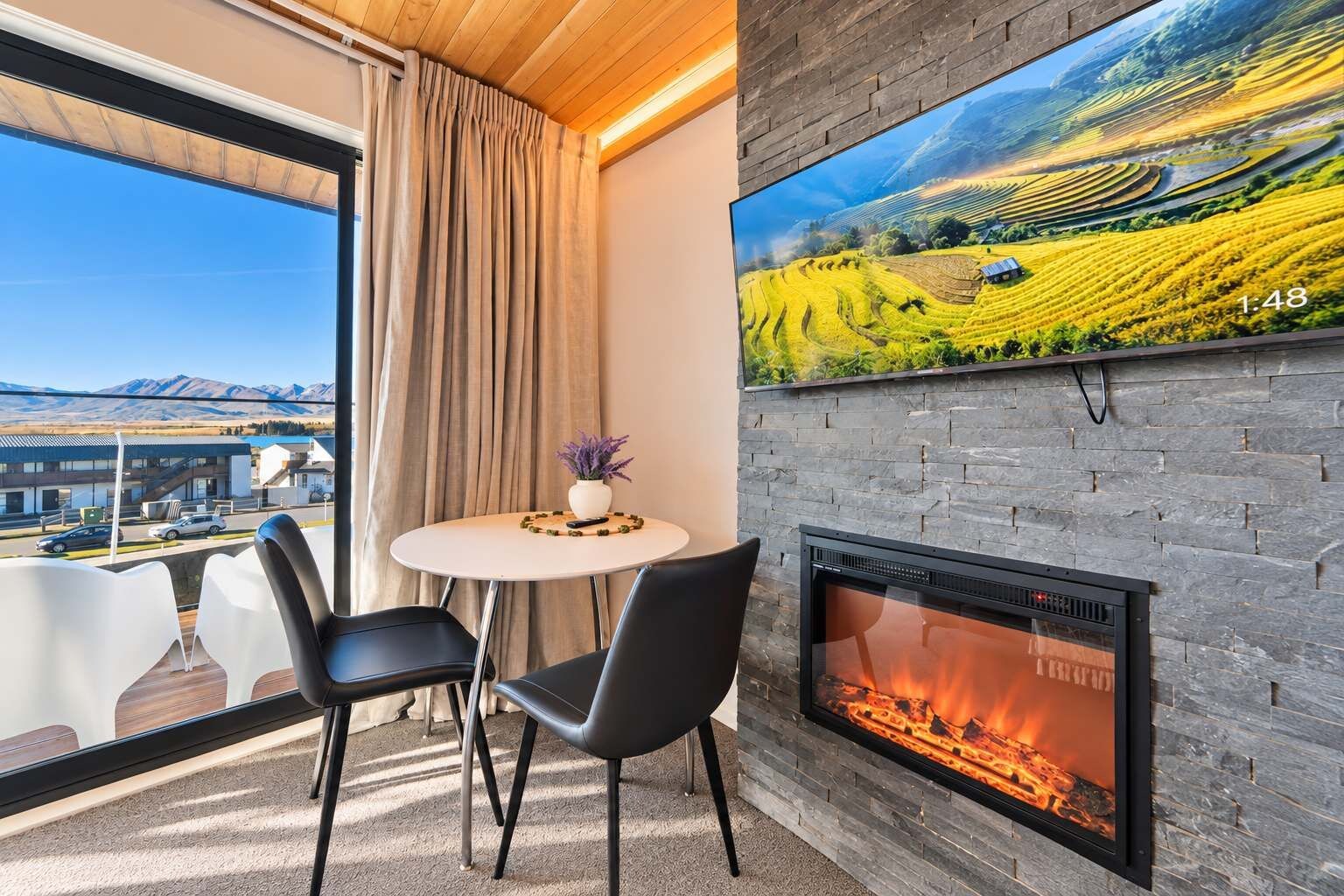 A modern room with a fireplace, a round table with two chairs, a large window with a view of mountains, and a painting of a landscape.