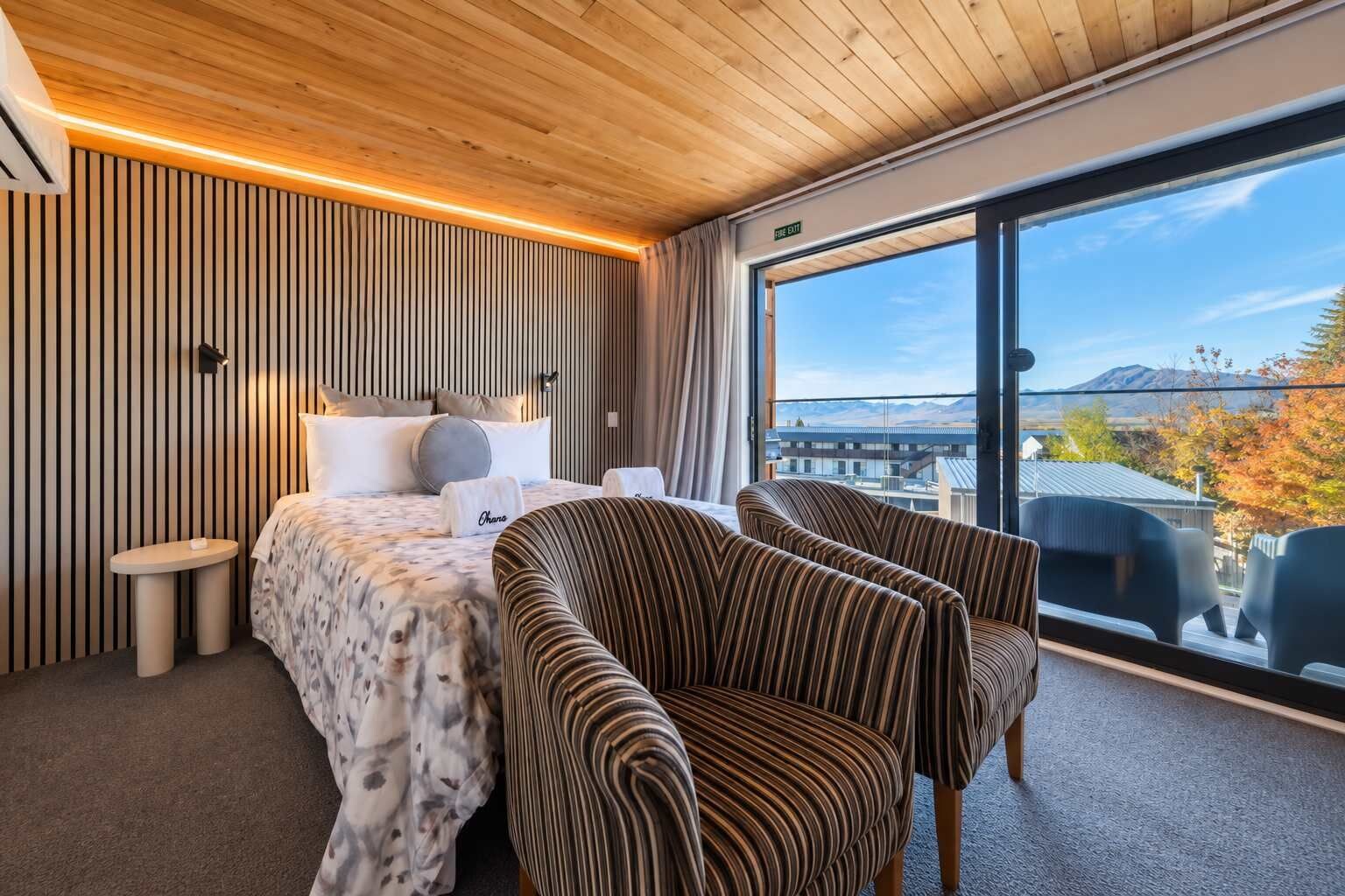 A modern hotel room with a large bed, two striped armchairs, a small table, and a view of the cityscape through large windows.