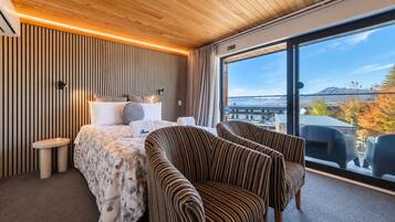 A modern hotel room with a large bed, two striped armchairs, a small table, and a view of the cityscape through large windows.