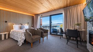 A modern hotel room with a bed, two striped armchairs, a dining table with chairs, a fireplace, and a large window with a view. - Starry Retreat | Lake Views + Fireplace (Lake Tekapo)