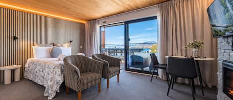 Una habitación de hotel moderna con una cama, dos sillones a rayas, una mesa de comedor con sillas, una chimenea y un ventanal con vistas.