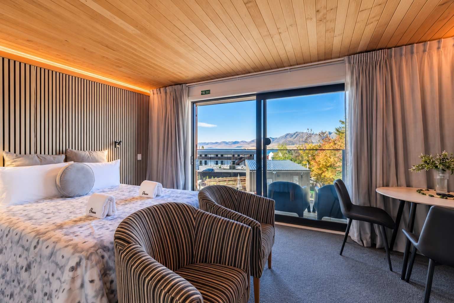 A hotel room with a bed, two striped armchairs, a small table, and a view of mountains through large windows.