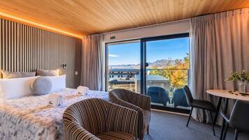 A hotel room with a bed, two striped armchairs, a small table, and a view of mountains through large windows.