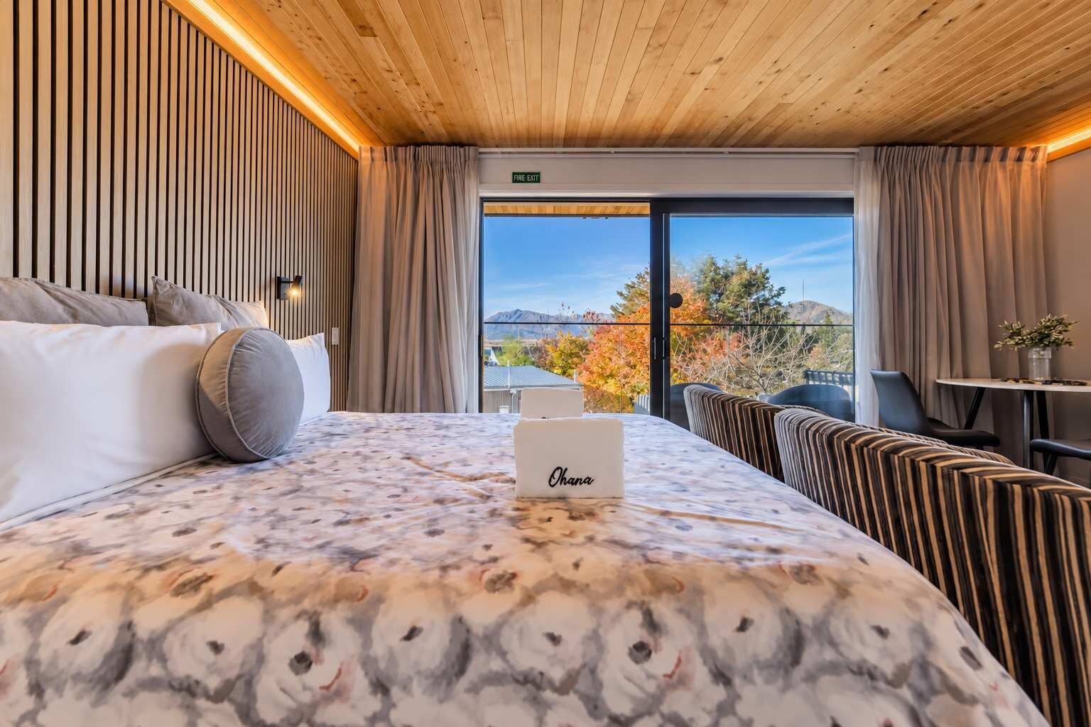Quarto de hotel com cama, mesa de cabeceira, poltrona, sofá, janela com vista para árvores e montanhas, e placa de saída de incêndio.
