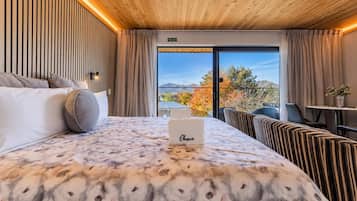 Quarto de hotel com cama, mesa de cabeceira, poltrona, sofá, janela com vista para árvores e montanhas, e placa de saída de incêndio.