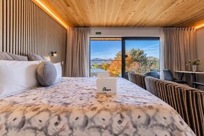 A hotel room with a bed, a bedside table, a chair, a sofa, a window with a view of trees and mountains, and a fire exit sign. - Starry Retreat | Lake Views + Fireplace (Lake Tekapo)