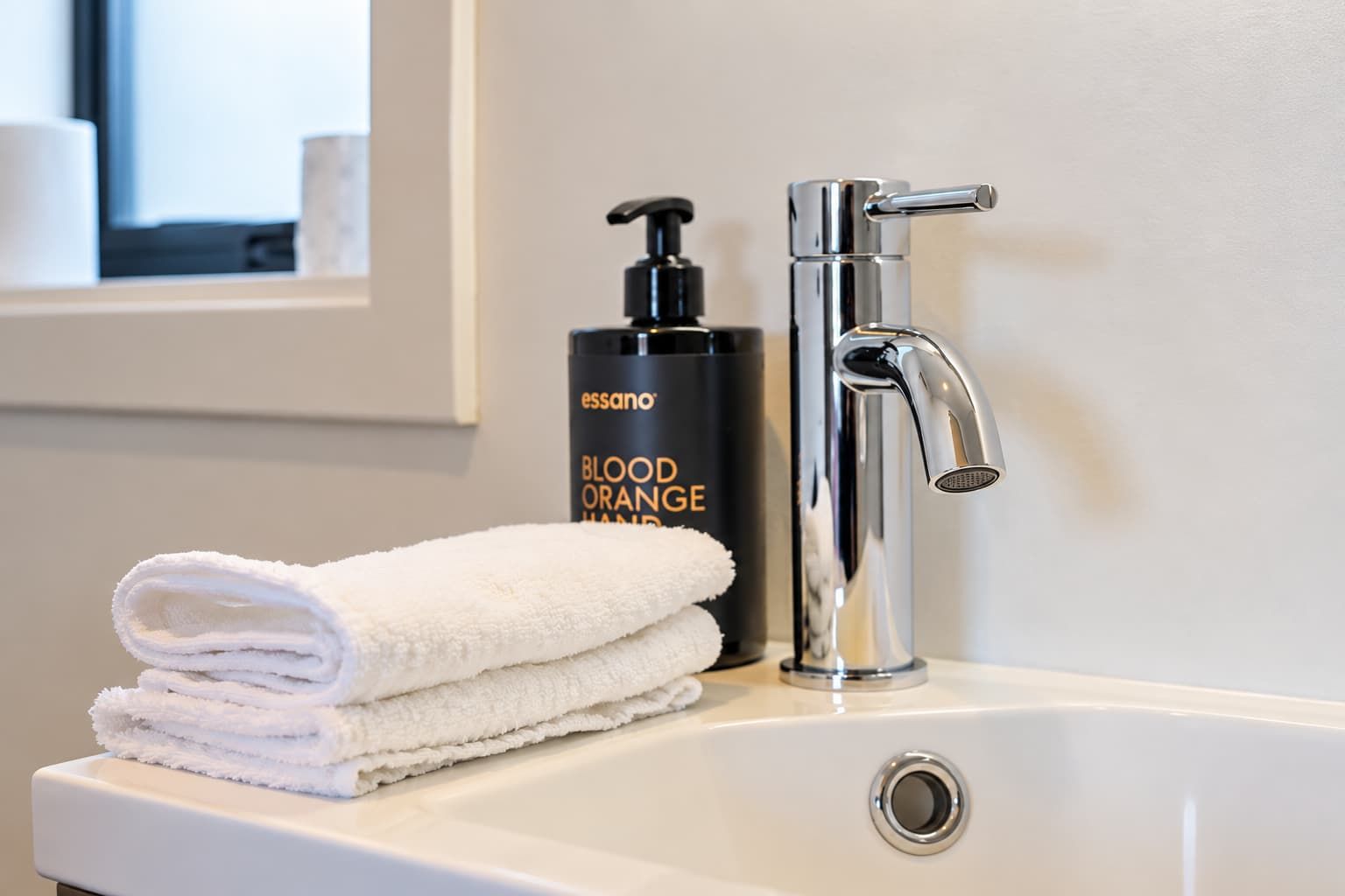 Un lavamanos de baño con un grifo moderno, una botella de jabón de manos Essano Naranja Sanguínea y una pila de toallas blancas.
