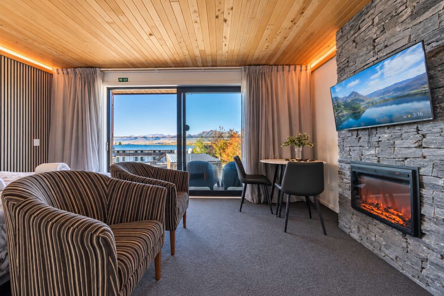 A hotel room with a fireplace, a dining area, and a large window with a view of mountains and a lake.