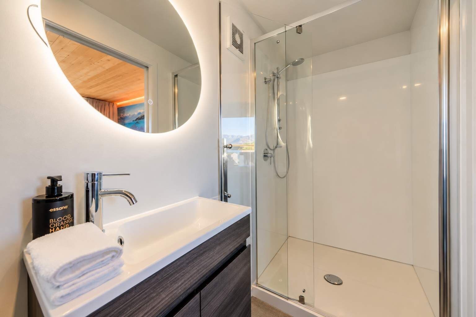 A modern bathroom with a glass-enclosed shower, a white sink with a dark cabinet, and a round mirror with a wooden ceiling view.