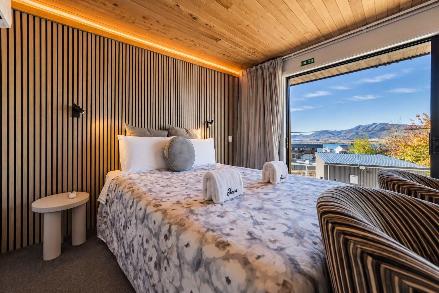 A hotel room with a bed, a bedside table, a chair, and a view of the outdoors.