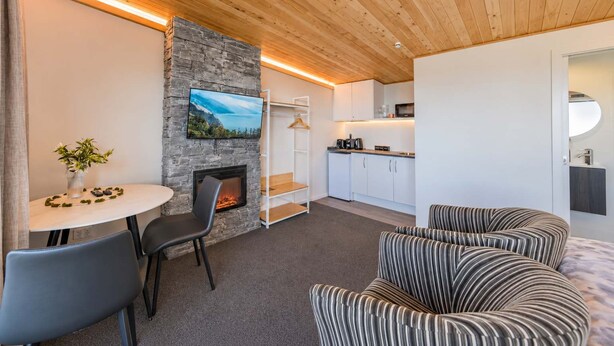 A cozy living area with a fireplace, a television mounted on a stone wall, a dining table with chairs, and a striped sofa. - Starry Retreat | Lake Views + Fireplace (Lake Tekapo)