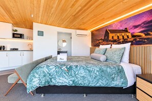 A modern bedroom with a bed, bedside table, and a view of a building through a window. - Starlight Escape | Lake Views + Balcony (Lake Tekapo)