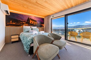 A hotel room with a bed, two chairs, a nightstand, a large window with a view, and a mural on the wall. - Starlight Escape | Lake Views + Balcony (Lake Tekapo)