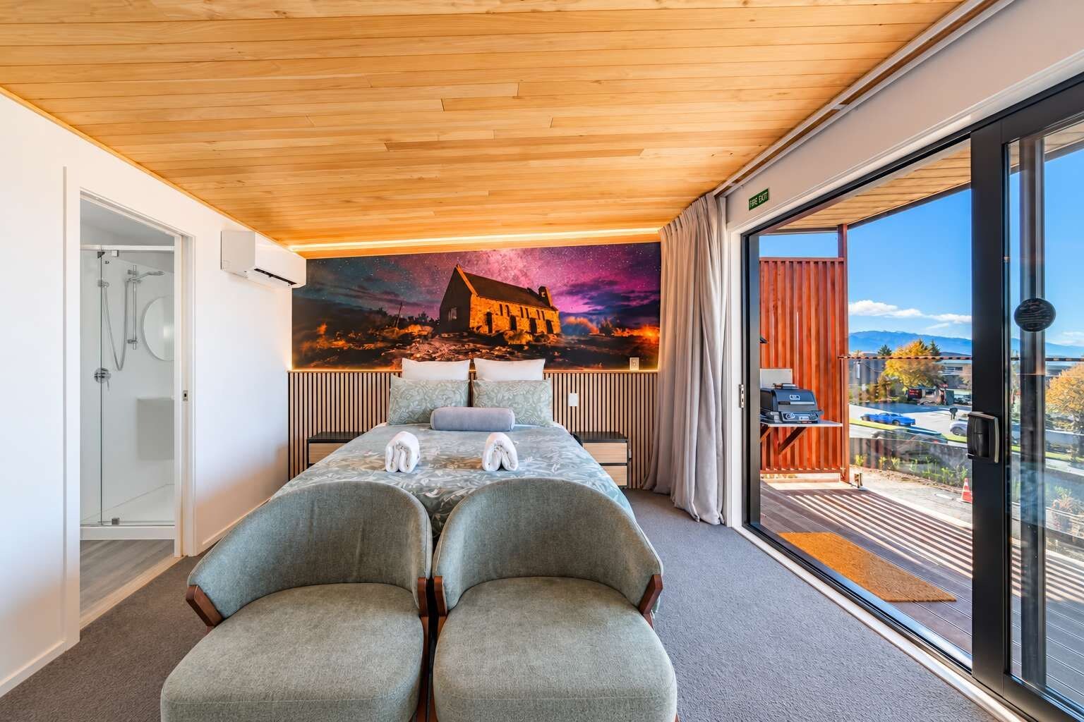 Habitación de hotel con cama, dos sillones, ducha, ventana grande con vistas y un mural en la pared.