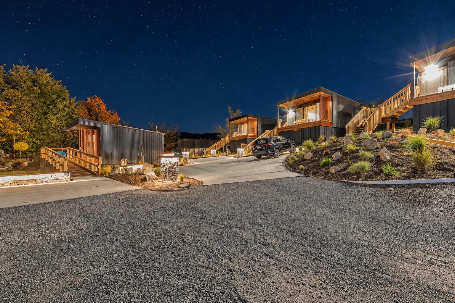 A modern residential area with multiple houses connected by a pathway, featuring a gravel driveway and landscaped areas.