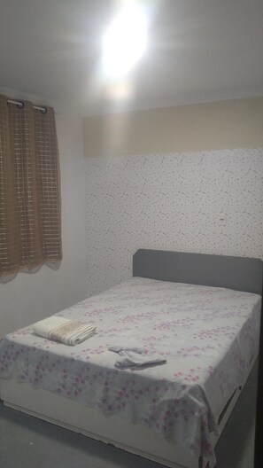 Ein kleines Zimmer mit einem Einzelbett, einer schlichten, floralen Tagesdecke, einer Wand mit dezentem Muster und einem braunen Vorhang am Fenster.