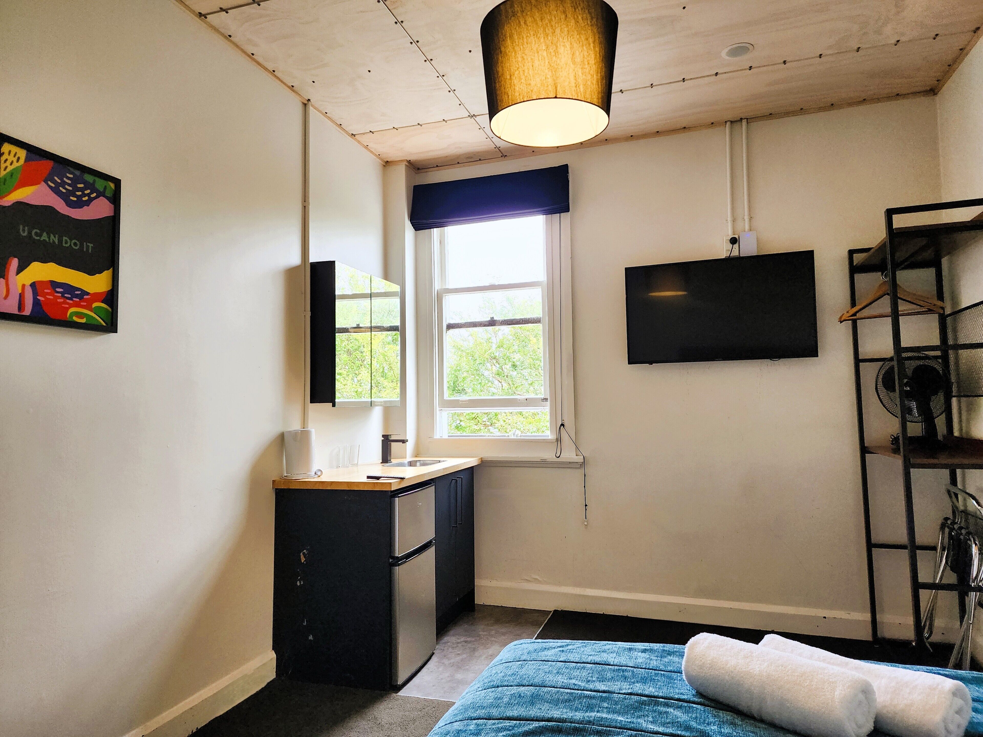 Una habitación pequeña y bien iluminada con kitchenette, una ventana con vistas, un televisor montado y una cama con cubierta azul.