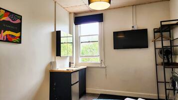 Una habitación pequeña y bien iluminada con kitchenette, una ventana con vistas, un televisor montado y una cama con cubierta azul.
