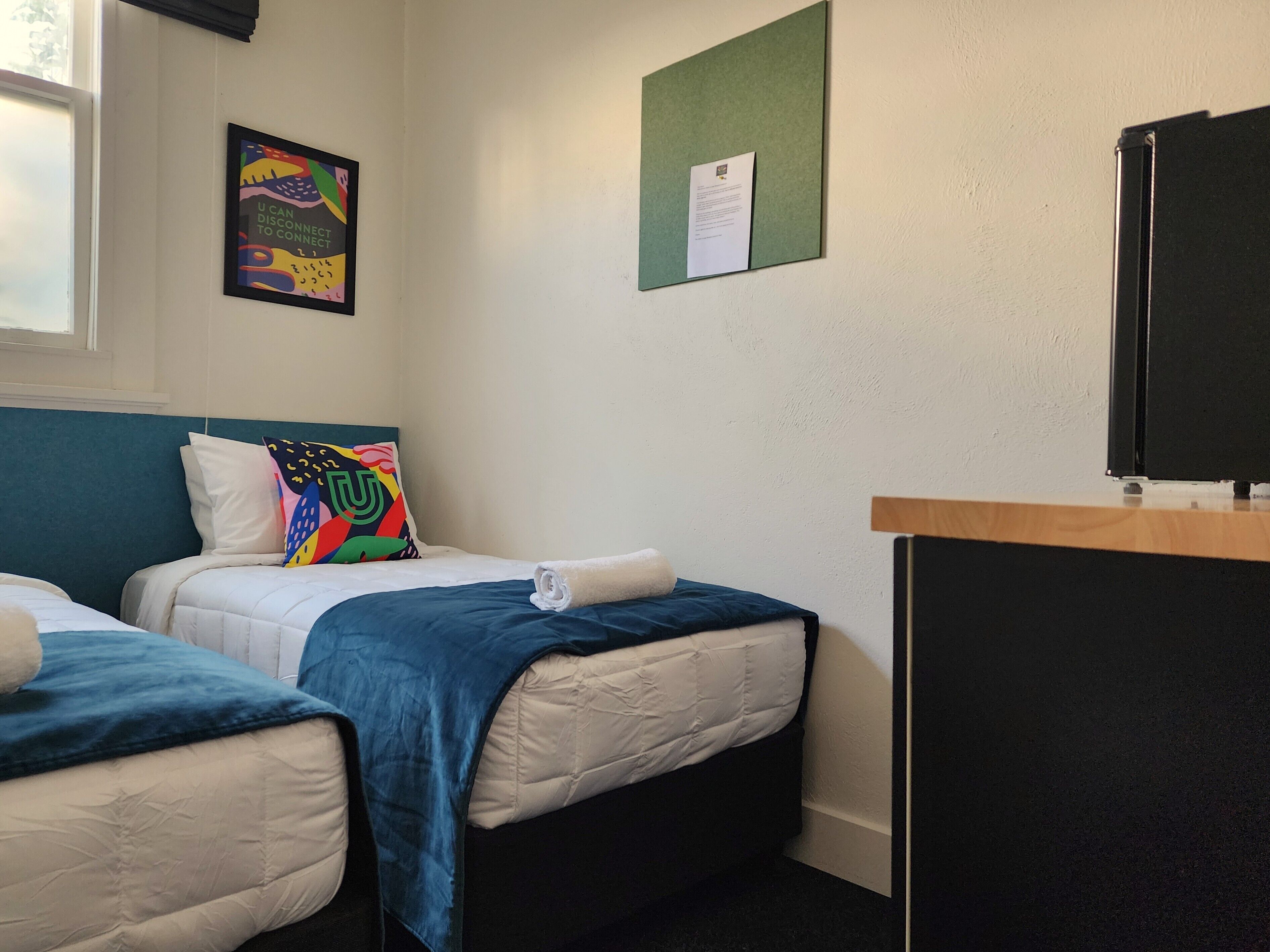A hotel room with two beds, a desk, a bulletin board, and a window.