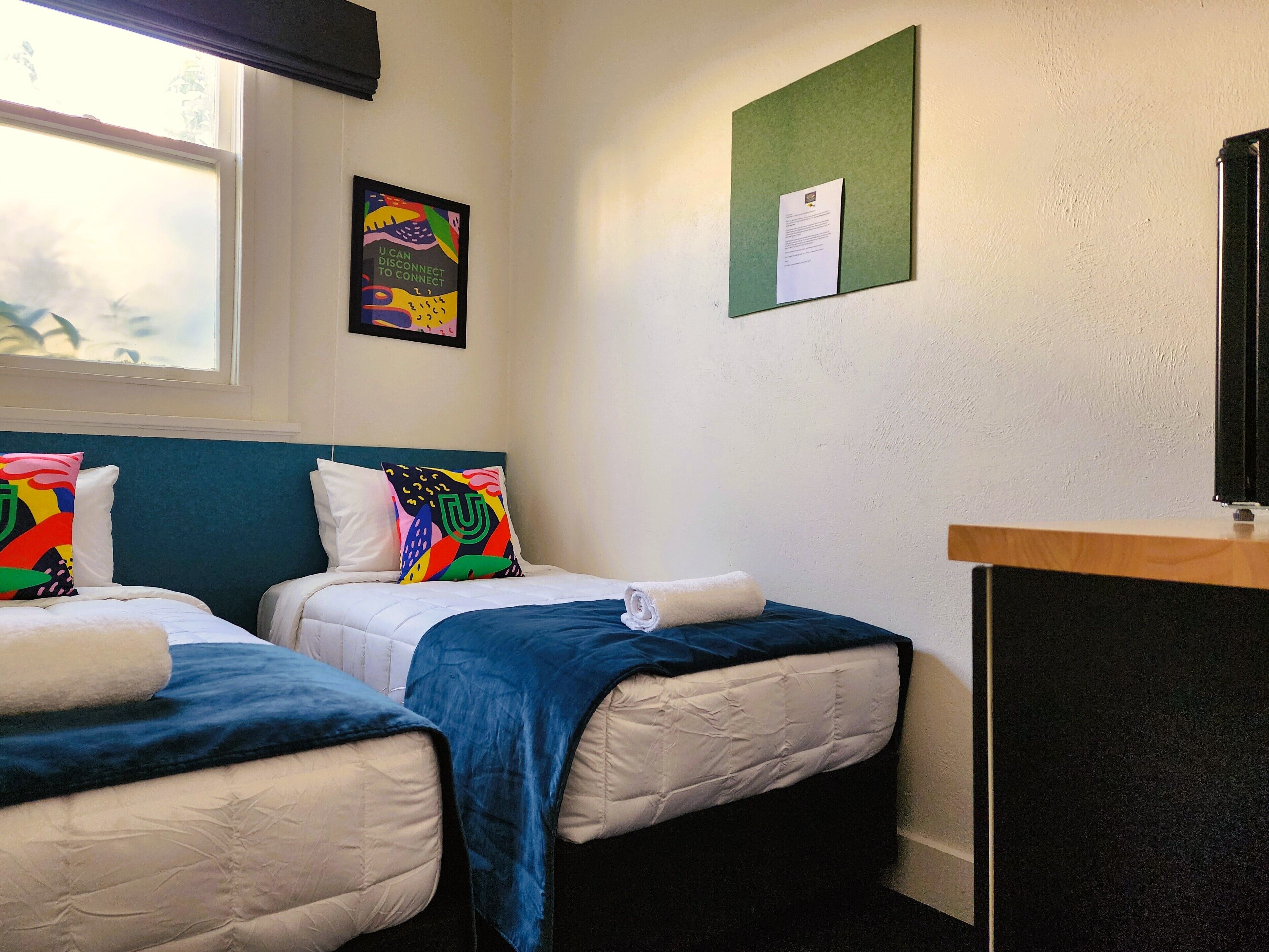 A hotel room with two beds, a window, a bulletin board, and a desk.