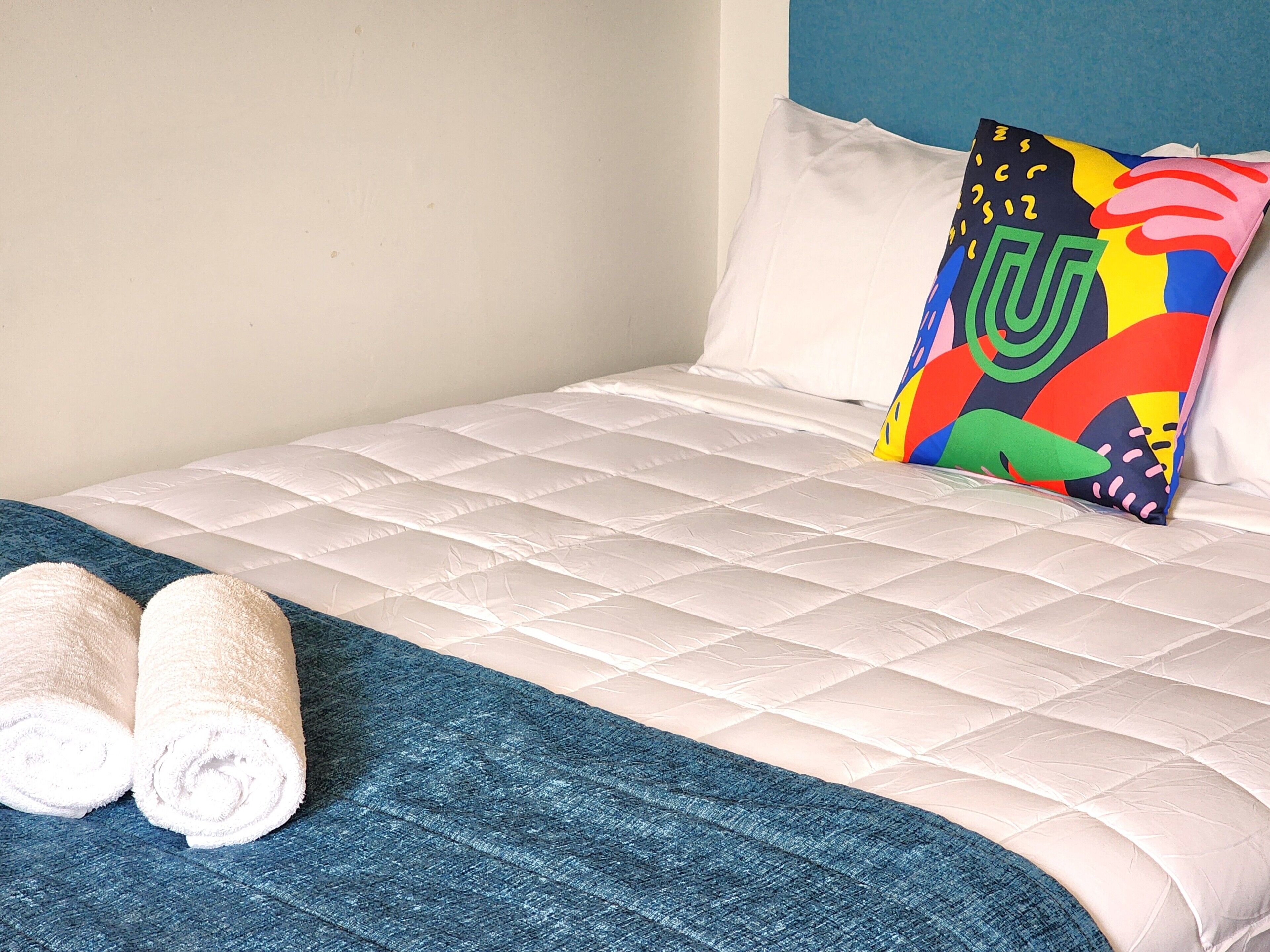 A neatly made bed with a quilted white duvet, a colorful pillow, and rolled white towels on a blue bedspread.