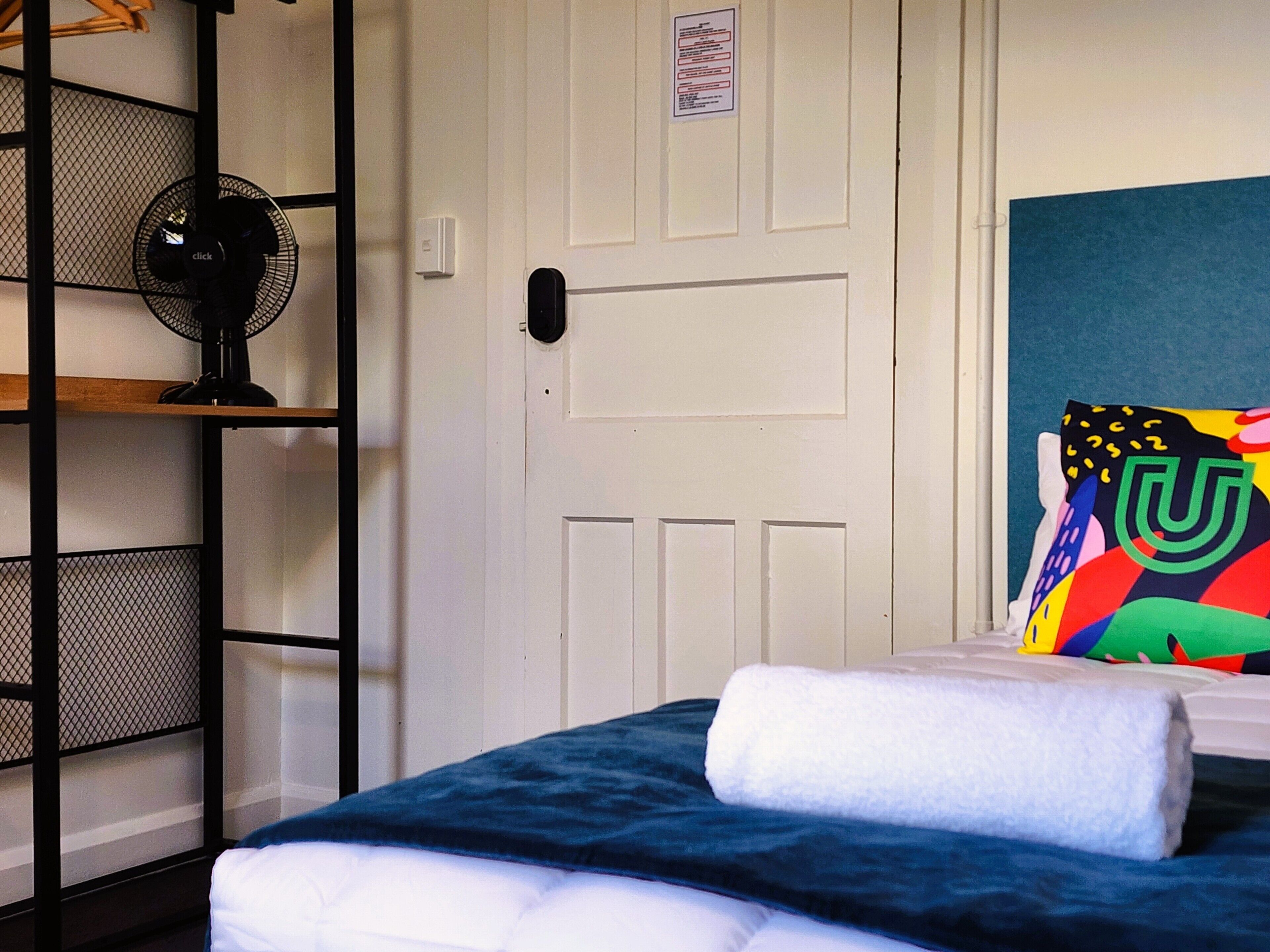 Una habitación con una cama, una puerta blanca, un ventilador negro y un cojín colorido.