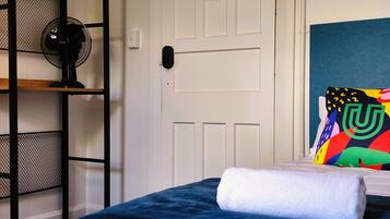 Una habitación con una cama, una puerta blanca, un ventilador negro y un cojín colorido.
