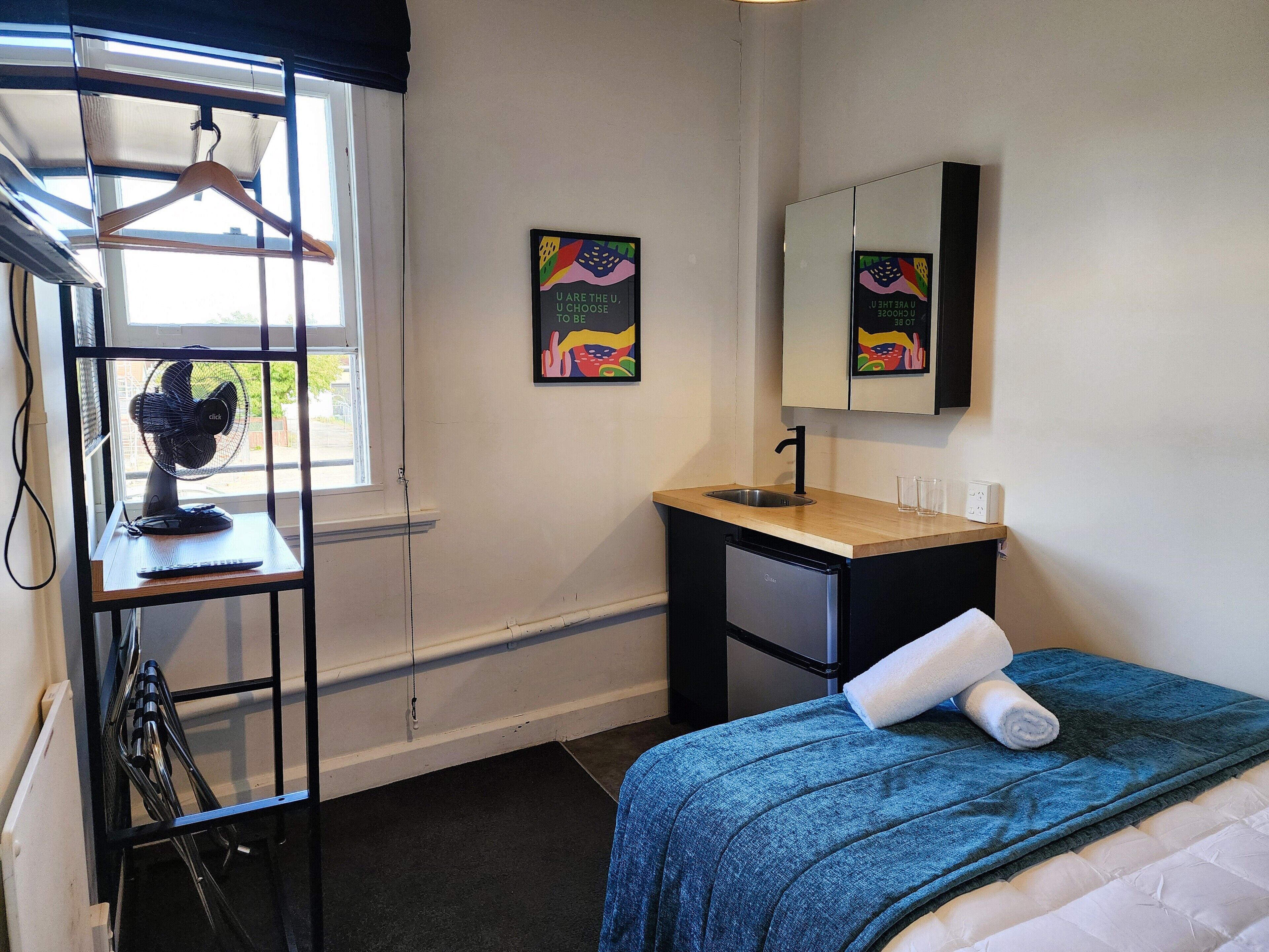 A bedroom with a bed, a desk with a sink, a mirror, and a window with a fan.