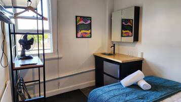 A bedroom with a bed, a desk with a sink, a mirror, and a window with a fan.