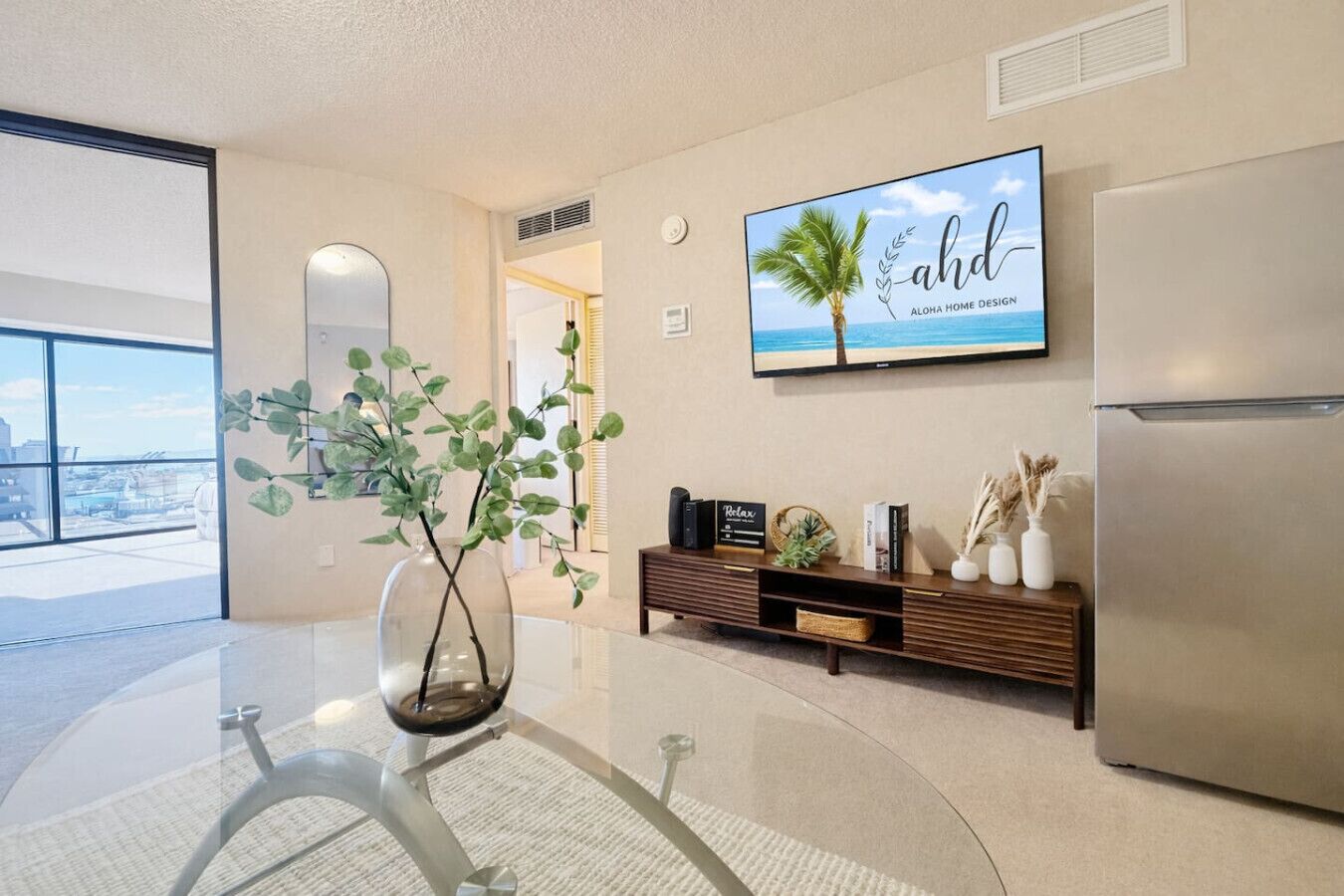 A modern living room with a glass table, a decorative plant, a wall-mounted TV, and a built-in refrigerator.