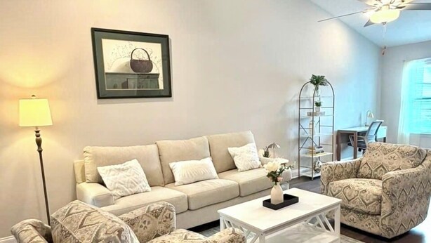 Un salon comprenant un canapé, deux fauteuils à motifs, une table basse, une lampe sur pied, un ventilateur de plafond et un tableau encadré accroché au mur.
