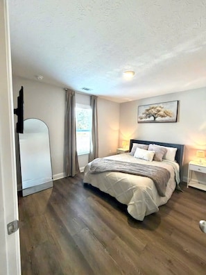 Une chambre à coucher avec un lit, une table de chevet, un tableau et une fenêtre avec des rideaux.
