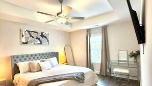 Une chambre à coucher comprenant un lit, un ventilateur de plafond, une table de chevet, un bureau avec une chaise et une fenêtre avec des rideaux.