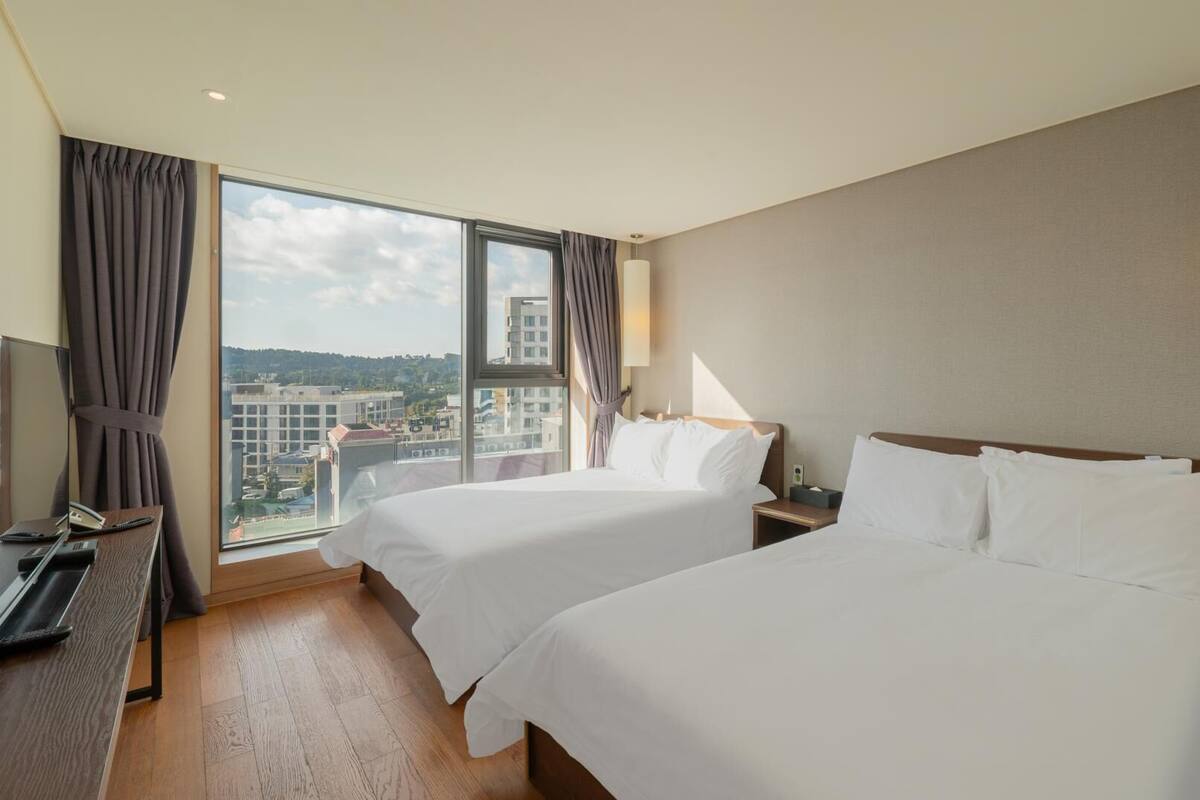 A hotel room with two beds, a TV, a desk, and a large window offering a city view.