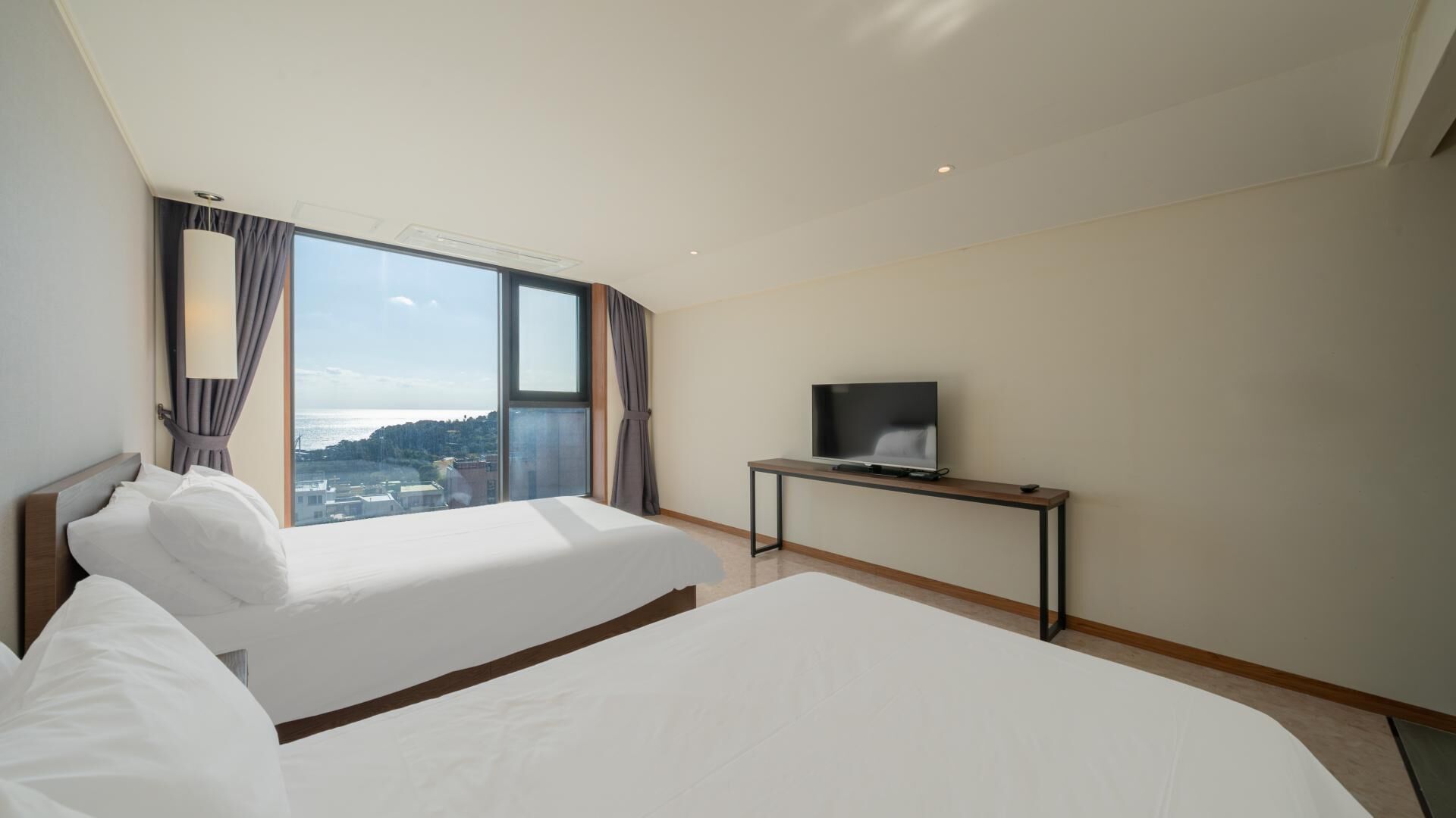 Habitación de hotel con dos camas, un televisor sobre un escritorio y un ventanal con vistas a la ciudad.