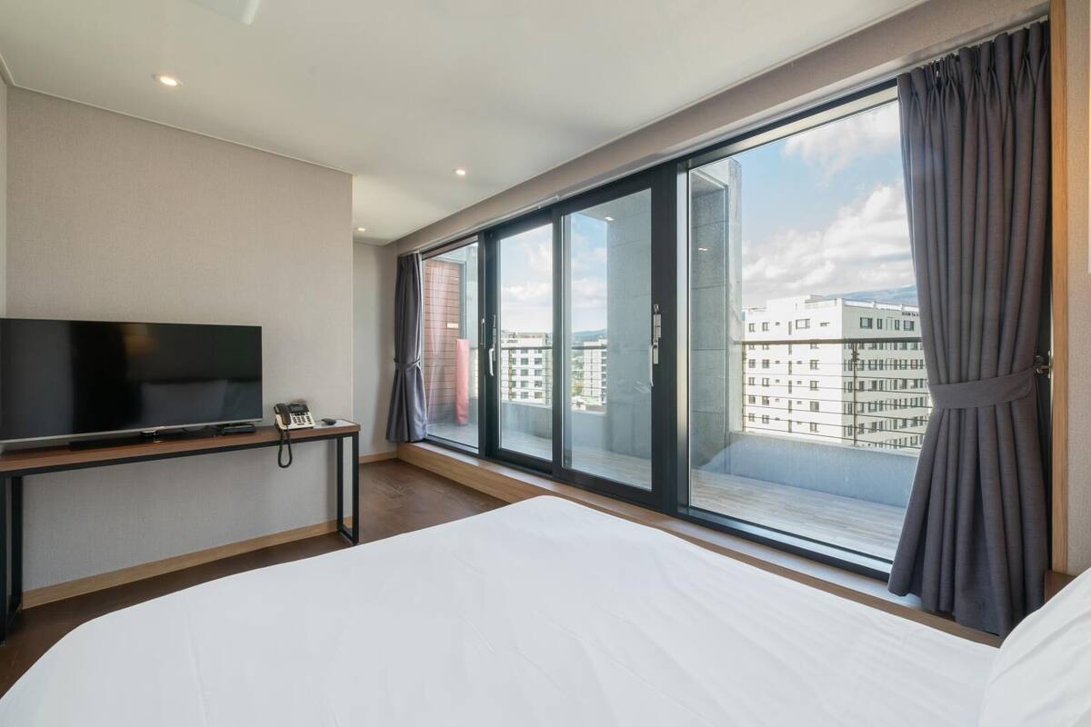 A hotel room with a large bed, a TV, a desk with a phone, and a view of buildings through large windows.