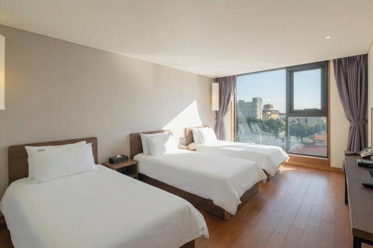A hotel room with two beds, a desk, a lamp, and a large window with a city view.