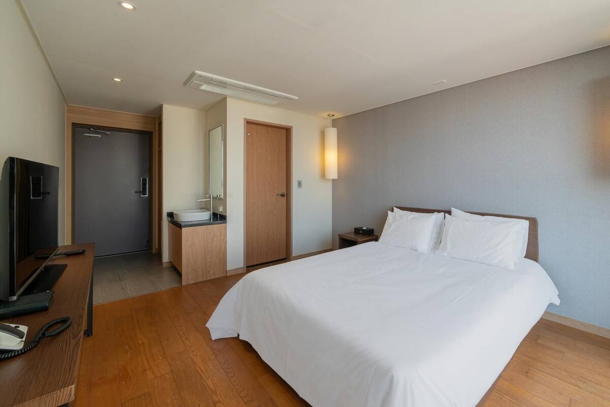 A hotel room with a bed, a desk with a computer, a TV, a bathroom with a sink and mirror, and a wall-mounted light fixture.