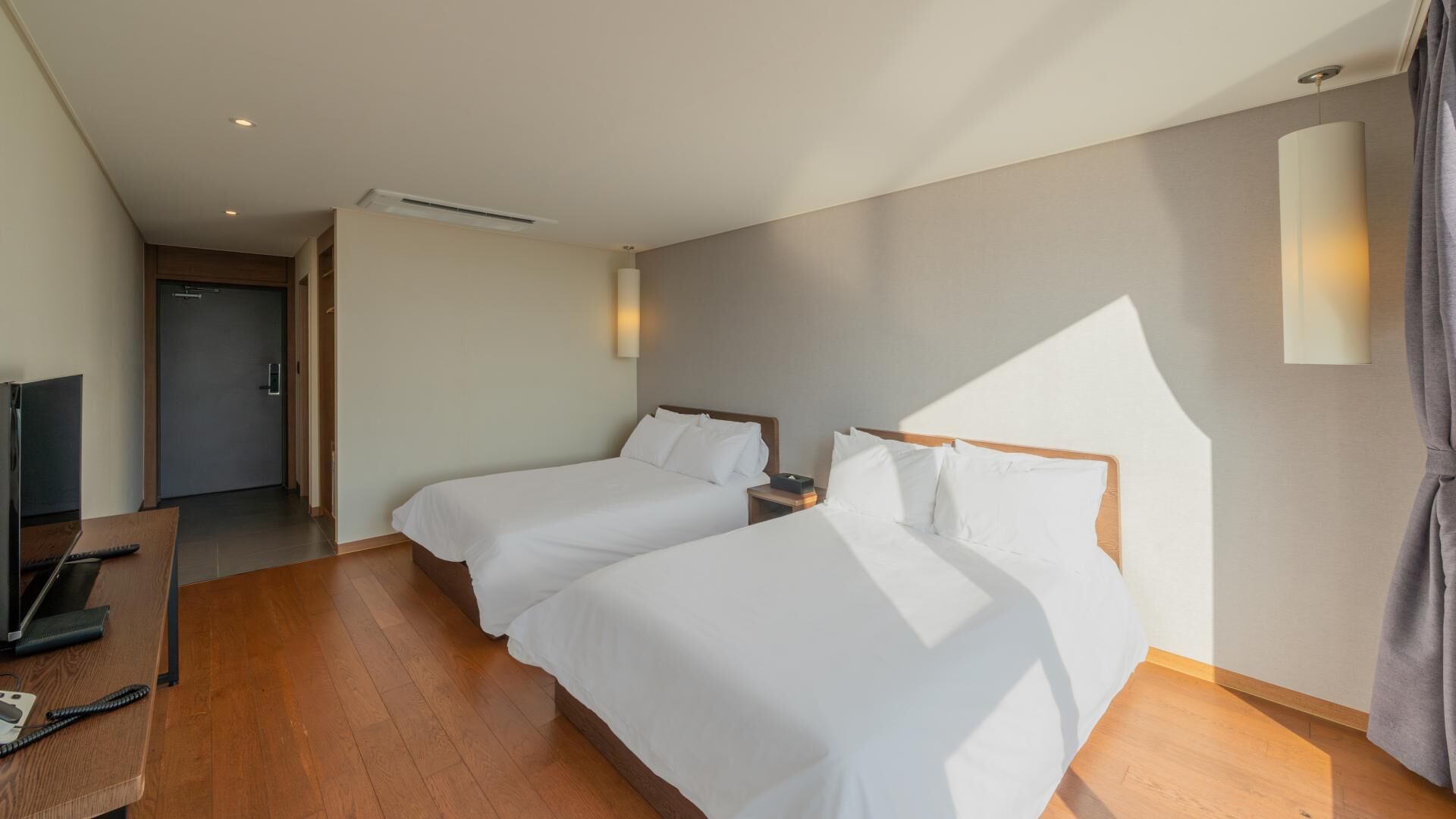 Habitación de hotel con dos camas, un escritorio con computadora, televisión y puerta que da a un pasillo.