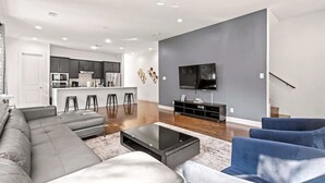 Een moderne woonkamer met een grijze hoekbank, een salontafel en blauwe fauteuils. Achtergrond toont een keuken met donkere kasten en een centraal eiland.