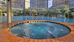 Una gran piscina exterior con una terraza de madera que la rodea, ubicada en un moderno complejo hotelero con varias edificaciones y palmeras.