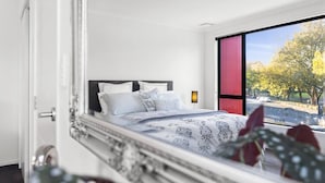Un dormitorio moderno con una cama grande, una ventana con vistas a árboles y una carretera, y una puerta que da a otra habitación.