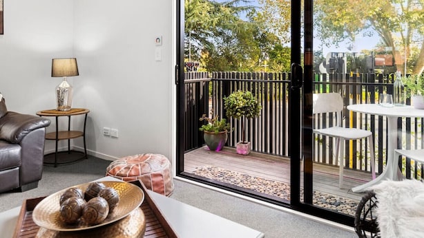 Una sala de estar moderna con un sofá, una mesita redonda con una lámpara, un cuenco decorativo y vistas a un balcón exterior con barandilla, plantas en macetas y una mesa con sillas.