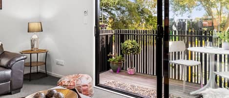 Un salon moderne comprenant un canapé, une petite table ronde avec une lampe, un vase décoratif, et donnant sur un balcon extérieur avec une balustrade, des plantes en pot et une table avec des chaises.