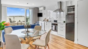 Cocina moderna con armarios blancos, estufa de acero inoxidable y horno empotrado. Hay una zona de comedor con mesa y sillas de vidrio, y un amplio ventanal con vistas a la ciudad.